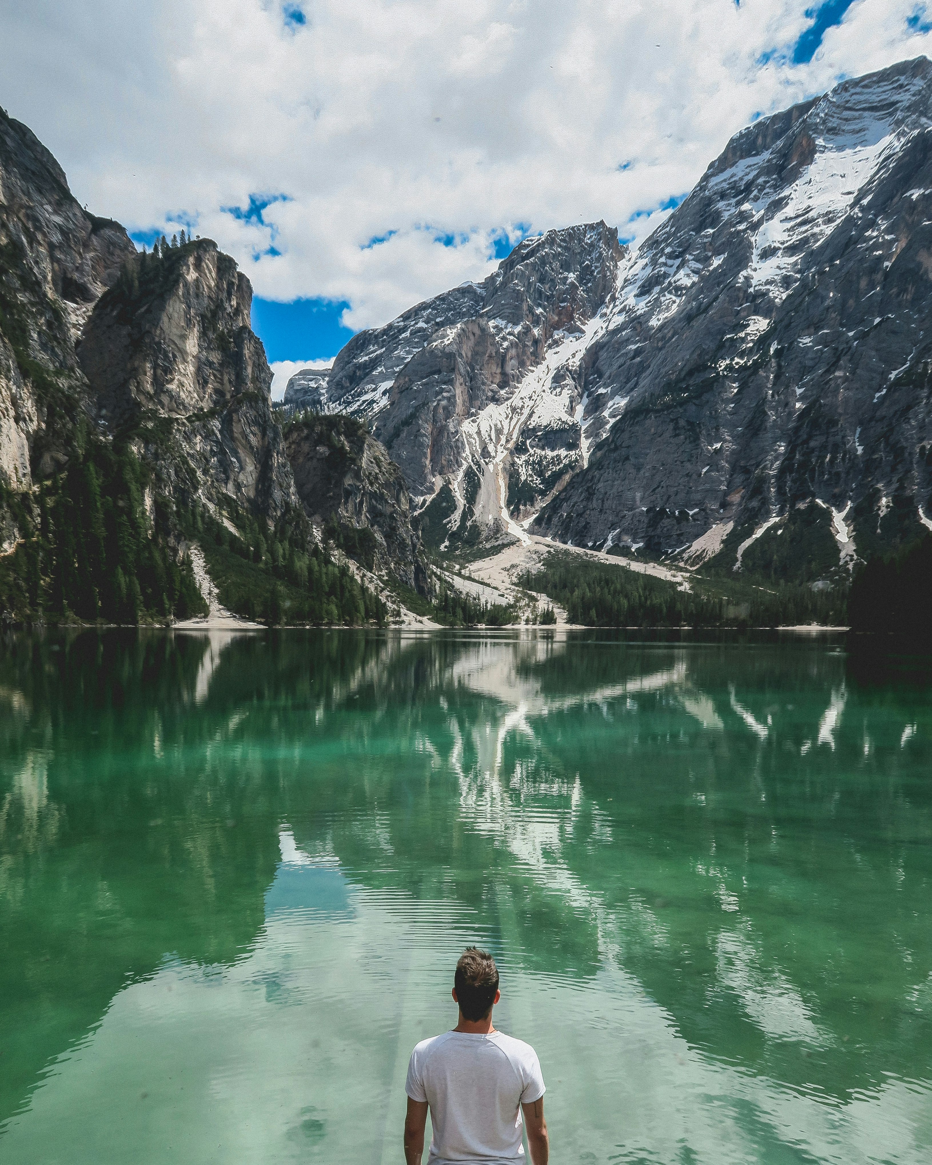 man standing in front of lake and cliff under white clouds and blue sky